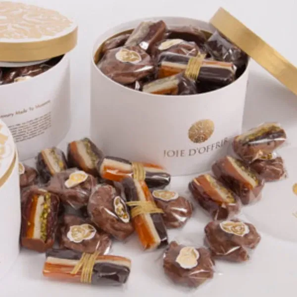 In the image, several elegant white boxes adorned with gold detailing are filled with assorted wrapped chocolates and candies, showcasing Joie d’Offrir's offerings in the Wedding Favours category. Each confection is individually wrapped with a decorative label, emphasizing attention to detail and quality. The boxes are partially open, revealing a variety of sweet treats, ideal for gifting to wedding guests as a memorable and tasteful token from the special day.