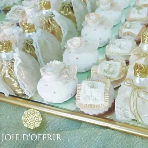 The image showcases an elegant display of wedding favors by Joie d’Offrir, categorized under Wedding Favours on the Mariage Elegant website in Canada. The favors, presented on a reflective gold tray, include beautifully crafted small boxes and pouches adorned with delicate lace, intricate ribbons, and ornate detailing. Some items feature floral embellishments, while others have personalized designs, suitable for a sophisticated wedding celebration.