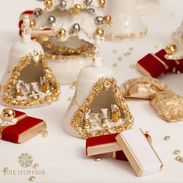 This image features elegant wedding favours by Joie d’Offrir, perfect for enhancing a sophisticated wedding aesthetic. The scene displays beautifully crafted ceramic bell ornaments, decorated with intricate gold detailing and miniature winter scenes inside. Accompanying these are stylishly wrapped gift boxes in rich red and gold tones, adding a touch of luxury. The decorative elements are arranged against a neutral background, subtly adorned with gold beads, highlighting the refined and celebratory ambiance ideal for weddings.