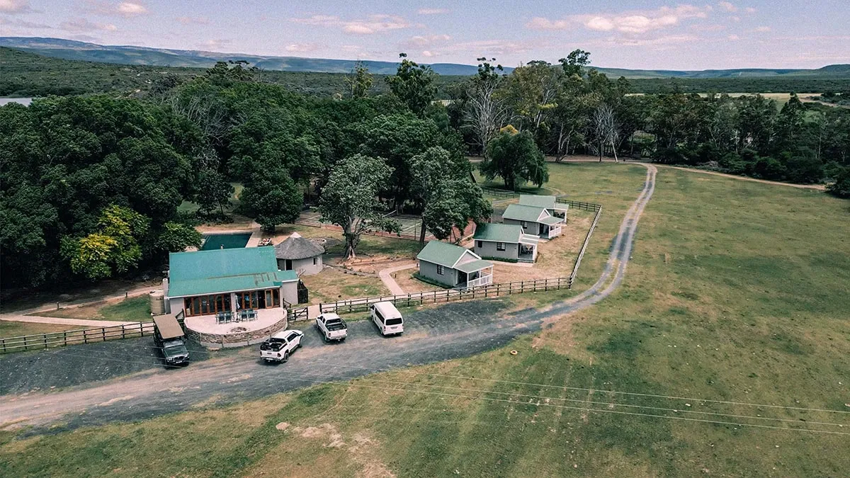 An aerial view of Glen Boyd Reserve, a picturesque destination wedding venue featured on Mariage Elegant, Canada. The image showcases several charming, rustic buildings with green roofs surrounded by lush greenery and expansive open fields. Multiple vehicles are parked along a narrow road leading to the venue, which is set against a backdrop of gently rolling hills and clear blue skies. The serene and natural setting offers an ideal location for a memorable wedding celebration.