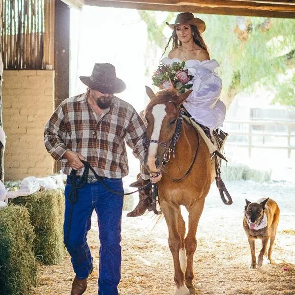 A bride wearing a white wedding dress and carrying a bouquet of flowers rides a horse led by a man in a plaid shirt and cowboy hat, symbolizing a rustic and adventurous destination wedding. A dog follows them, enhancing the countryside charm. This image highlights the unique and picturesque offerings of Glen Boyd Reserve, a destination wedding vendor featured on Mariage Elegant, Canada. The setting is filled with natural light, straw bales, and elements of a barn, creating an idyllic rural backdrop for a memorable wedding celebration.