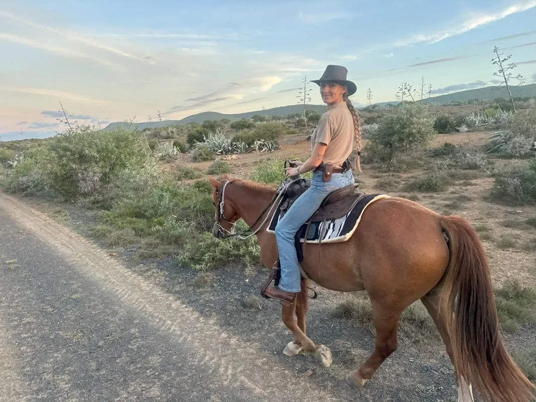 A person riding a brown horse on a dirt path at Glen Boyd Reserve, a destination wedding venue listed on Mariage Elegant in Canada. The rider is wearing a cowboy hat and casual attire, with a scenic backdrop of rolling hills and a partly cloudy sky. The setting reflects a rustic and adventurous theme suitable for unique destination weddings. The natural landscape surrounding the path enhances the venue's appeal for couples seeking an outdoorsy wedding experience.