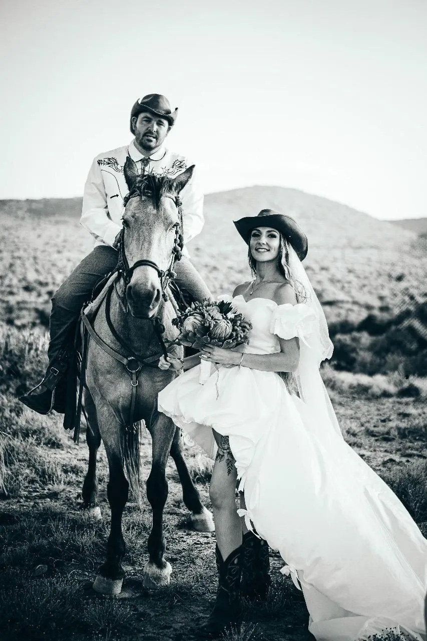 A bride in a flowing white gown and cowboy boots stands beside a man on horseback, both wearing cowboy hats, against a scenic backdrop of open fields and distant hills. The groom is seated on a brown horse adorned with Western-style gear. This picturesque scene captures the essence of a rustic destination wedding at Glen Boyd Reserve, highlighting the unique blend of elegance and country charm.