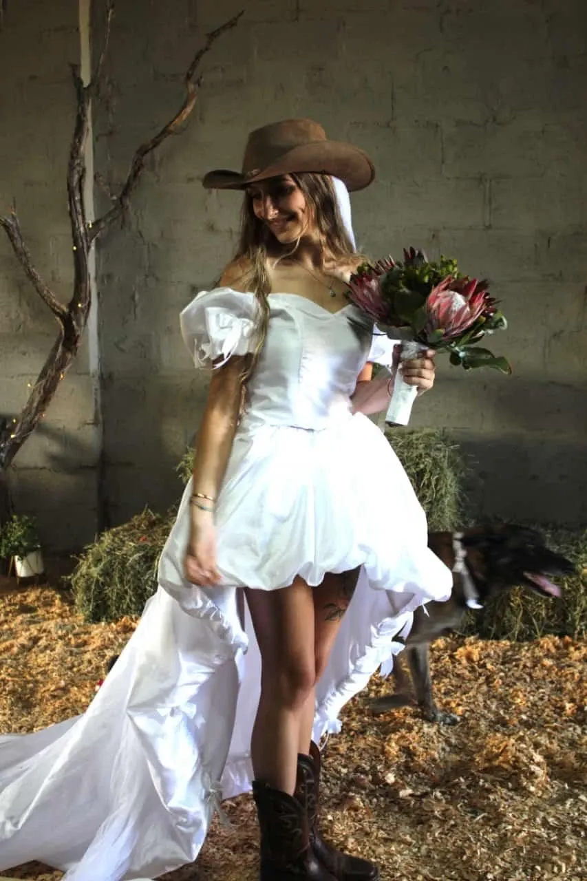 A bride in a unique, rustic setting wears a white off-shoulder wedding dress with a high-low hemline, complemented by a cowboy hat and boots, embodying a country chic style. She holds a bouquet of native flowers, reflecting a natural theme. The background features a barn-like interior with wooden accents and greenery, highlighting the ambiance suitable for a destination wedding by Glen Boyd Reserve. A dog is faintly visible in the background, adding a personal touch to the scene.