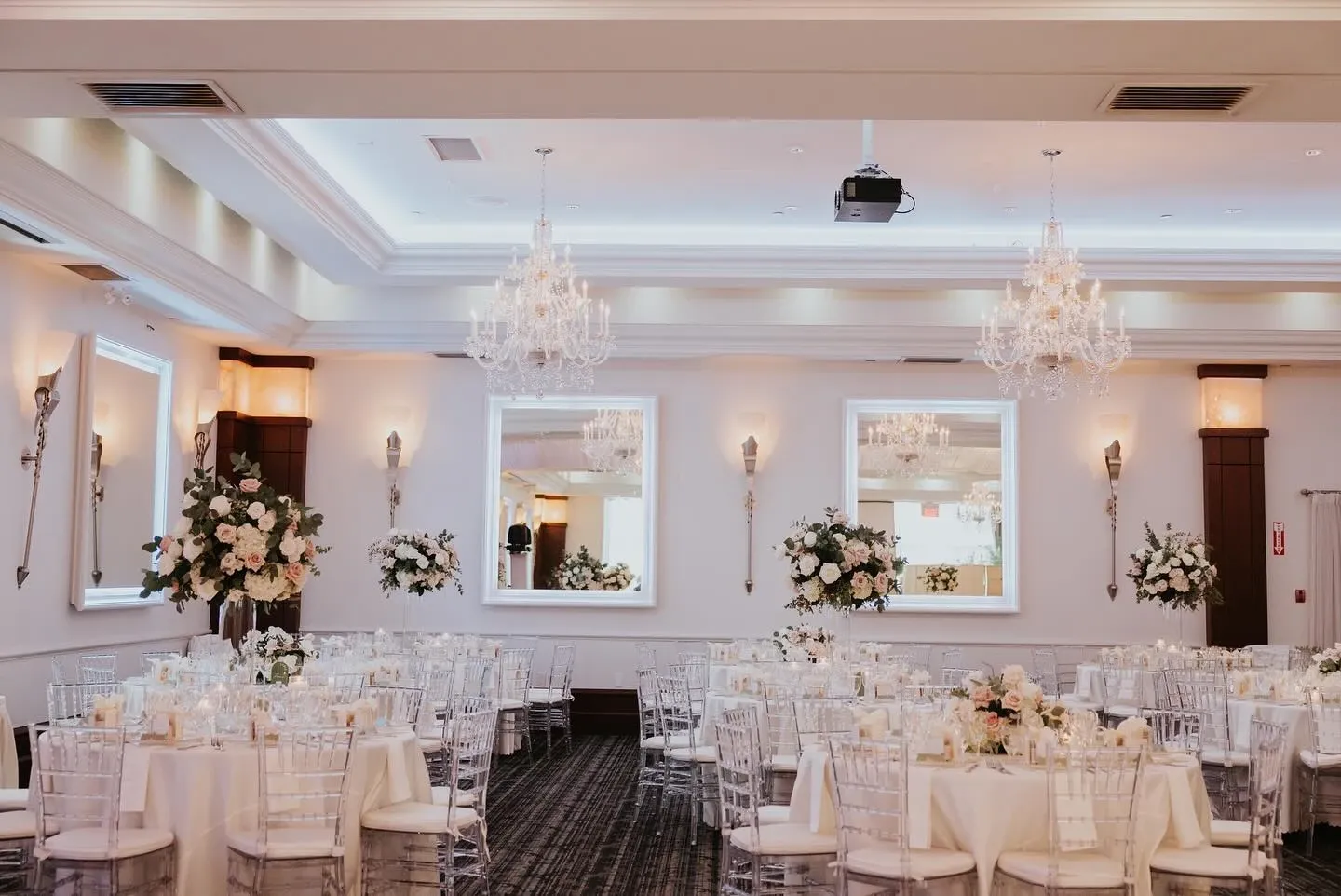 The image showcases a beautifully decorated wedding reception hall curated by Events by Paige Ligris, a notable vendor in the Wedding Planners category. The elegant setting features round tables draped in white tablecloths, each adorned with glowing candles and floral centerpieces primarily consisting of white and cream flowers. Crystal-clear, Chiavari chairs offer a sophisticated seating arrangement. The room is illuminated by grand chandeliers, enhancing the luxurious atmosphere. Large mirrors on the walls reflect the tasteful décor, making the space feel even more expansive and bright. This exquisite setup highlights the attention to detail and style offered by Events by Paige Ligris, making it ideal for those planning a wedding in Canada.