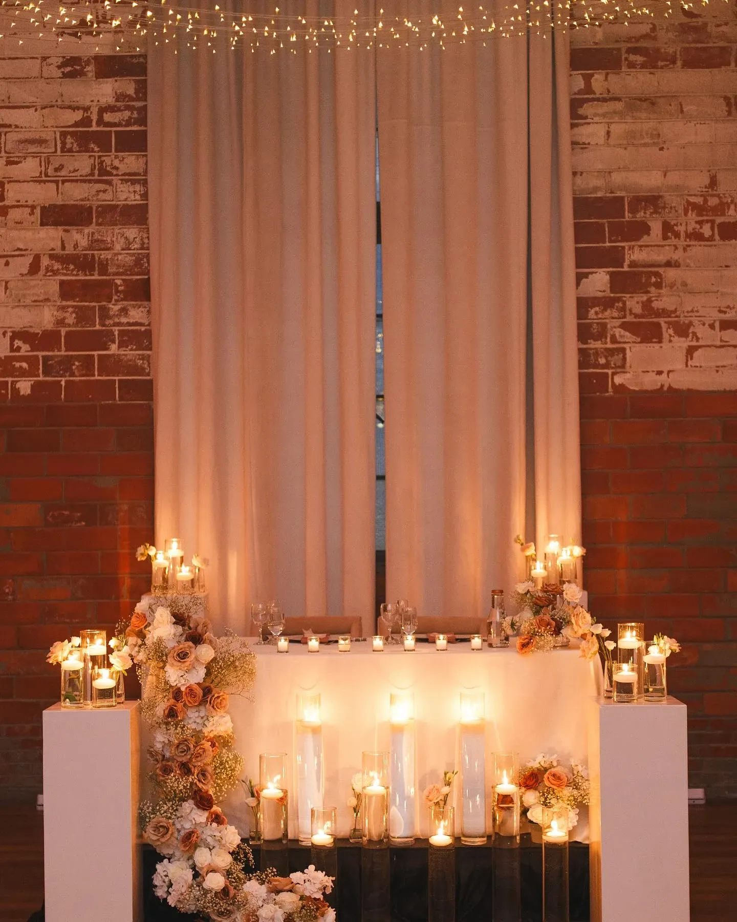 The image features a beautifully decorated wedding altar by Events by Paige Ligris, a renowned wedding planner in the Mariage Elegant vendor listing. The setting showcases a rustic brick wall backdrop with soft, beige curtains. The altar table is adorned with an array of candle arrangements, exuding a warm, romantic glow. Delicate floral bouquets in shades of white, peach, and blush elegantly frame the table, enhancing the romantic ambiance. Above, fairy lights create a whimsical atmosphere, perfect for an intimate wedding ceremony.