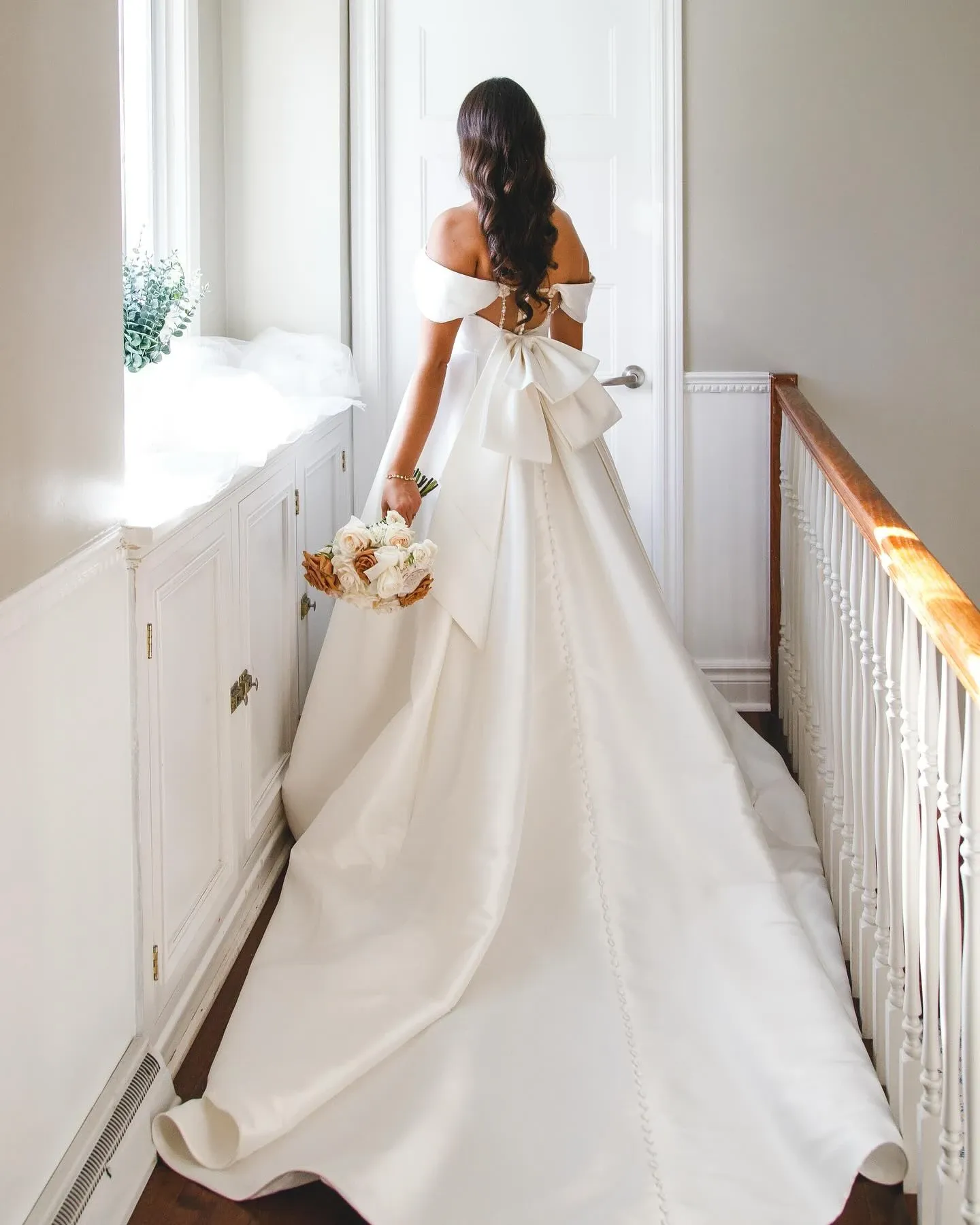 In this elegant image from Mariage Elegant's wedding planning section, a bride in a luxurious, off-the-shoulder white gown stands at the top of a staircase. The gown features an elaborate bow at the back, complemented by a long, flowing train adorned with buttons. She holds a bouquet of white and peach flowers, contributing to the soft and romantic ambiance. The bright, softly lit hallway enhances the romantic setting, ideal for showcasing the expertise of Events by Paige Ligris in the Wedding Planners category.