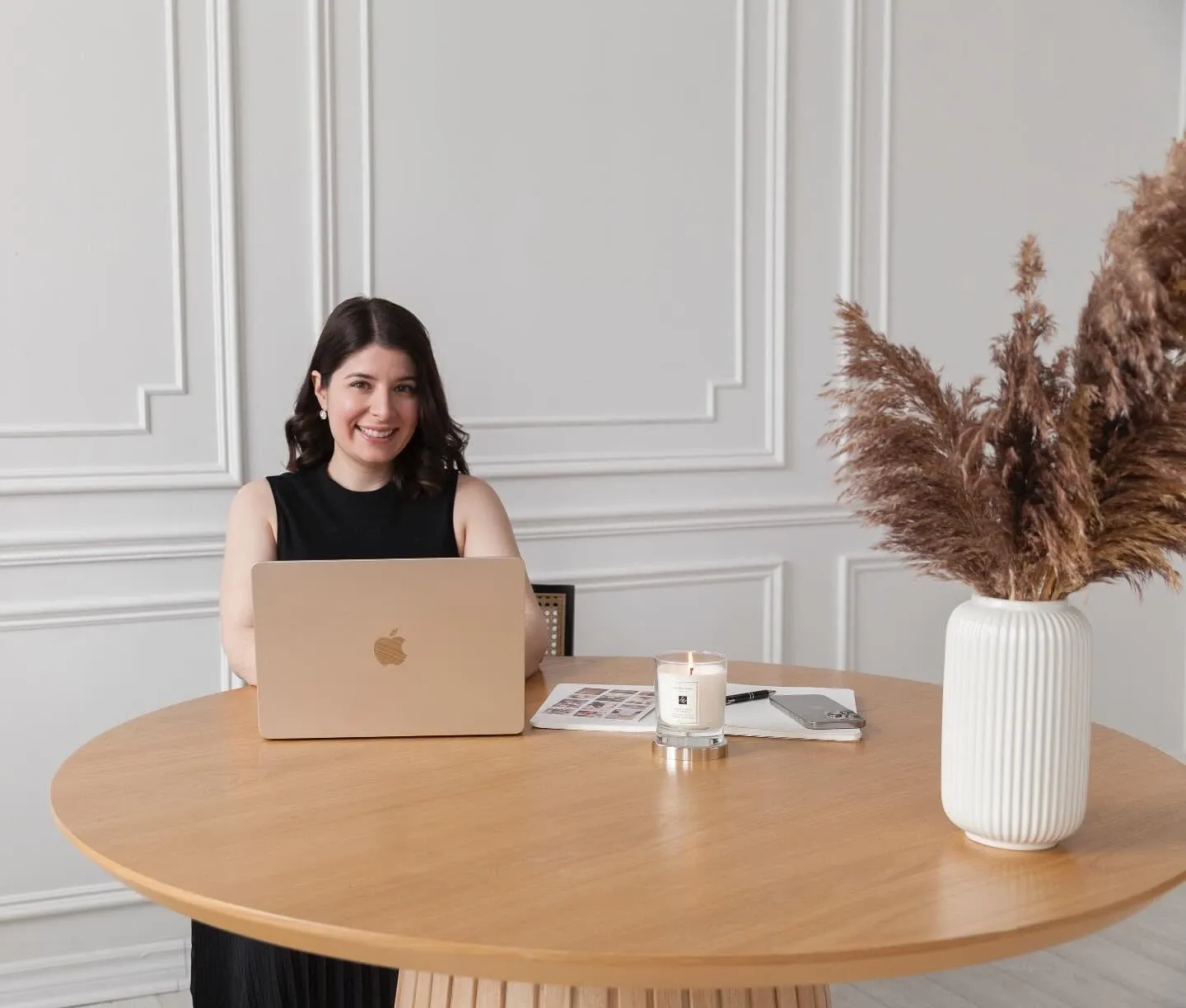 A woman is seated at a round, light wood table using a laptop, with a gentle smile that conveys professionalism. She appears to be working, possibly planning wedding details as part of her role with Events by Paige Ligris, a wedding planner in the Mariage Elegant vendor listing. The room features elegant, white wainscoted walls, enhancing the sophisticated atmosphere. On the table, there is a lit candle, wedding planning brochures, and a notepad, which suggest a focus on creating memorable wedding experiences. A vase filled with pampas grass sits on the table, adding a touch of rustic elegance.