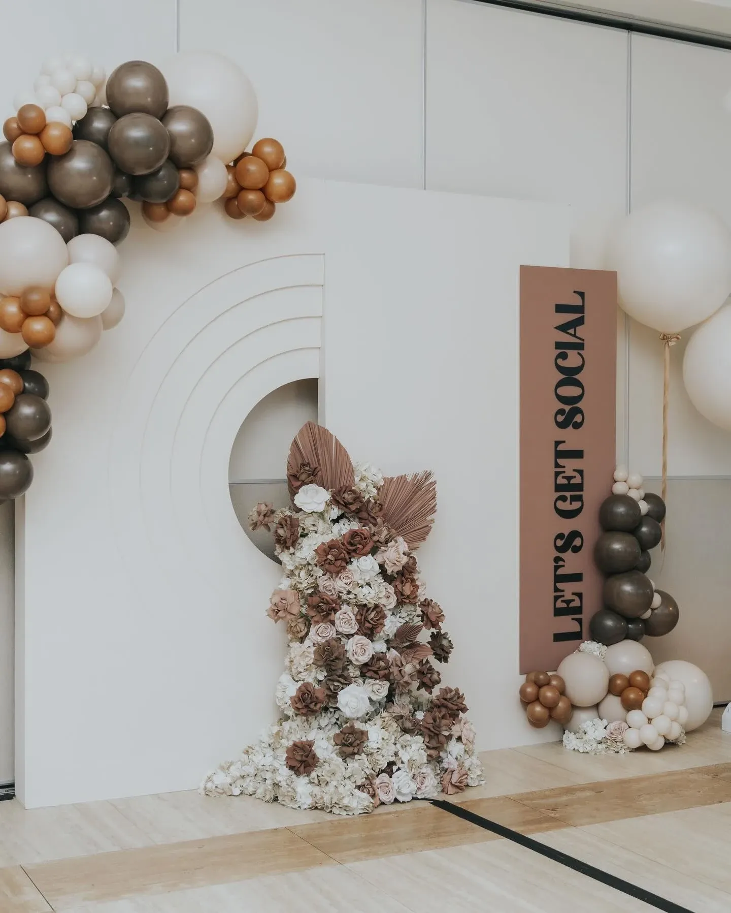 This image showcases a stylish event setup by Events by Paige Ligris, categorized under Wedding Planners. It features a white backdrop with a modern arch design adorned with an elegant arrangement of balloons in neutral tones, including beige, brown, and cream. At the base of the arch, there is a beautiful floral display composed of roses and other blooms in soft pink, white, and deep reddish-brown hues. A tall sign with the phrase 'Let's Get Social' adds a contemporary touch to the decor. This setup is ideal for a chic, sophisticated wedding atmosphere, illustrating the attention to detail offered by Events by Paige Ligris.