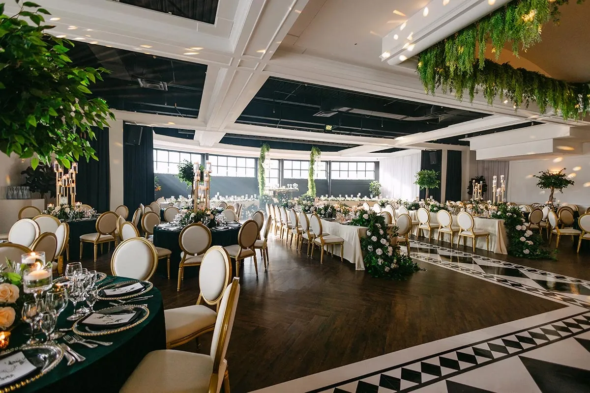 The image showcases a beautifully decorated wedding venue arranged by Events by Paige Ligris, a vendor specializing in wedding planners. The setting features elegantly set round and rectangular tables adorned with floral arrangements in soft colors, complemented by lush greenery hanging from the ceiling. The floor displays an intricate black and white geometric pattern, adding sophistication to the atmosphere. Comfortable white chairs with gold trim surround the tables, and candlelit centerpieces enhance the romantic ambiance. The open design with large windows allows natural light to fill the space, creating an inviting and elegant environment perfect for a wedding celebration.