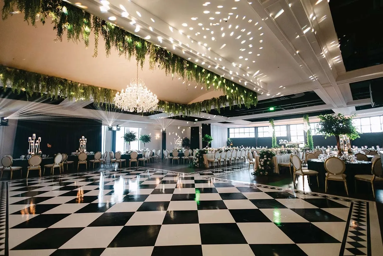 The image showcases a beautifully decorated wedding venue planned by Events by Paige Ligris, categorized under Wedding Planners. The venue features an elegant black and white checkered dance floor, which is the central focus of the room. Overhead, a stunning crystal chandelier adds a touch of glamour, complemented by hanging greenery that creates a lush atmosphere. The dining area is adorned with white chairs and tables set with fine glassware, ready for a sophisticated reception. Soft, ambient lighting enhances the romantic setting, highlighting the intricate details and exquisite decor typical of a wedding planned by a top-tier vendor in Canada.