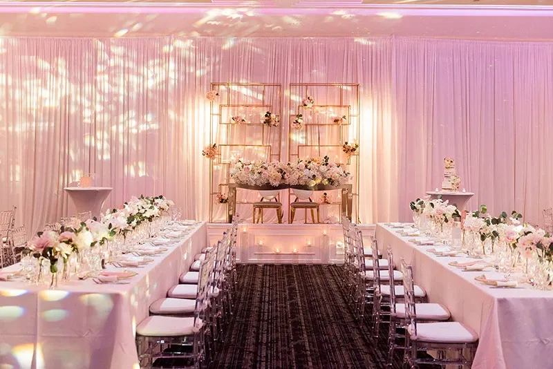 The image shows a beautifully decorated wedding reception area set up by Events by Paige Ligris, categorized under Wedding Planners. The venue is elegantly arranged with long tables draped in white tablecloths, adorned with delicate floral centerpieces featuring pink and white flowers. Clear acrylic chairs are neatly aligned alongside the tables. At the front, there is a decorative backdrop with shelves displaying floral arrangements, adding a sophisticated touch. Soft, ambient lighting creates a warm and inviting atmosphere, reflecting off the textured white drapery that surrounds the room.