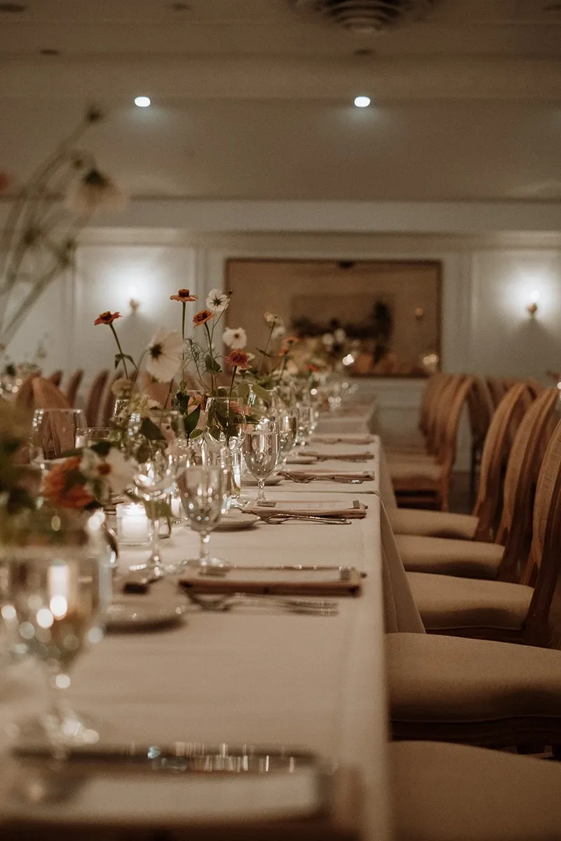 This image features a beautifully set wedding reception table by Events by Paige Ligris, a vendor specializing in wedding planning. The long table is elegantly adorned with a crisp white tablecloth and a series of delicate floral arrangements featuring small, colorful wildflowers. Crystal-clear wine glasses, silver cutlery, and neatly folded napkins are strategically placed, creating an inviting and sophisticated atmosphere. The venue's ambient lighting casts a warm, welcoming glow, enhancing the romantic setting typical of wedding celebrations. Ideal for inclusion in Mariage Elegant’s wedding planners category, this image showcases the meticulous attention to detail that Events by Paige Ligris brings to wedding arrangements in Canada.