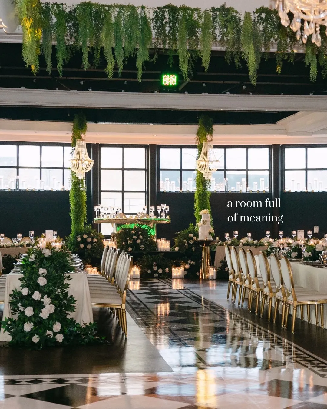 The image showcases an elegant wedding reception setup designed by Events by Paige Ligris, a prominent vendor in the Wedding Planners category. The venue is adorned with lush greenery cascading from the ceiling, creating a luxurious atmosphere. Long dining tables are elegantly set with white tablecloths and gold-trimmed chairs, surrounded by floral arrangements featuring white blooms and greenery. At the room’s center, a chic bar setup is visible, framed by additional floral decorations. Soft candlelight and sparkling glassware enhance the sophisticated ambiance, reflecting the high-standard wedding planning services offered by Paige Ligris in Canada.