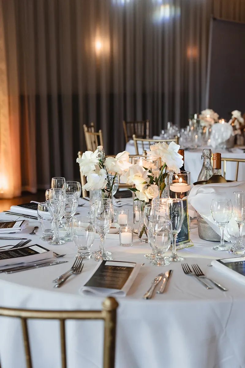 A beautifully set wedding reception table is elegantly arranged with delicate white floral centerpieces and surrounded by gold chiavari chairs. The table features neatly placed white plates, silver utensils, and crystal-clear glassware, emphasizing sophistication. Romantic candles are scattered across the table, casting a warm glow. A bottle of wine is chilling in an ice bucket, adding a touch of luxury. This image represents Events by Paige Ligris, a vendor in the Wedding Planners category on Mariage Elegant, Canada, highlighting their attention to detail and elegance in wedding setup.