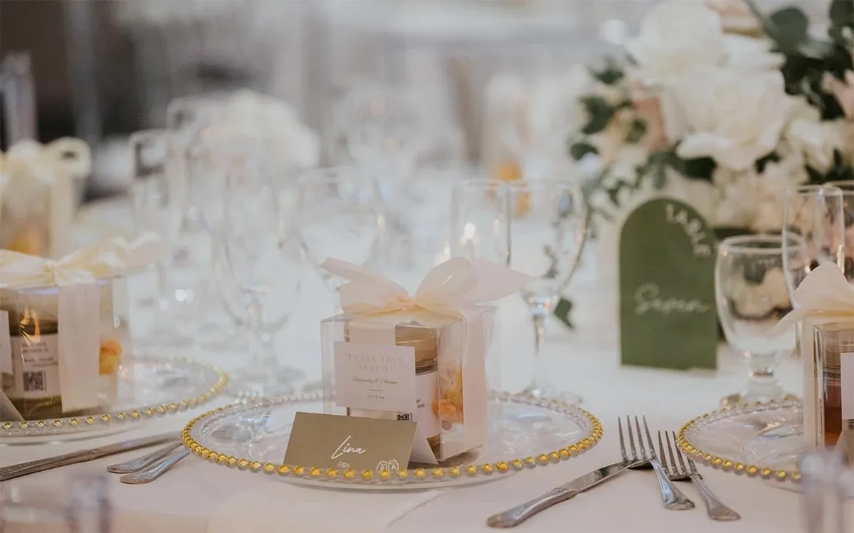 Elegant wedding table setting arranged by Events by Paige Ligris, a wedding planner in the Mariage Elegant vendor listing in Canada. The table features a sophisticated display with transparent glass plates decorated with gold beading, and an elegantly wrapped box on each setting. Delicate silverware is neatly placed on either side of the plates. Lush white floral arrangements, including roses and green foliage, serve as the table's centerpiece. A green place card, elegantly handwritten, enhances the arrangement, completing the luxurious wedding ambiance.