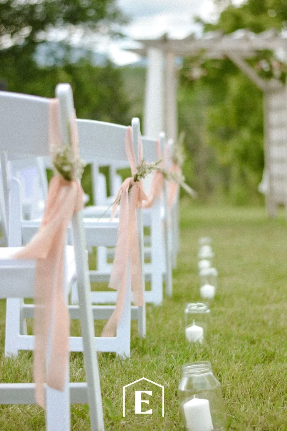 The image shows a beautiful outdoor wedding setup by Estrimont, categorized as a wedding venue. White wooden chairs are neatly arranged in rows on a lush green lawn, each chair adorned with soft pink ribbons and delicate sprigs of flowers, creating an elegant and inviting atmosphere. Aisle markers made of glass jars with white candles line the grassy path, leading to a wooden arbor draped with fabric in the background. The natural surroundings and thoughtful decorations suggest a serene and romantic setting ideal for a wedding ceremony.
