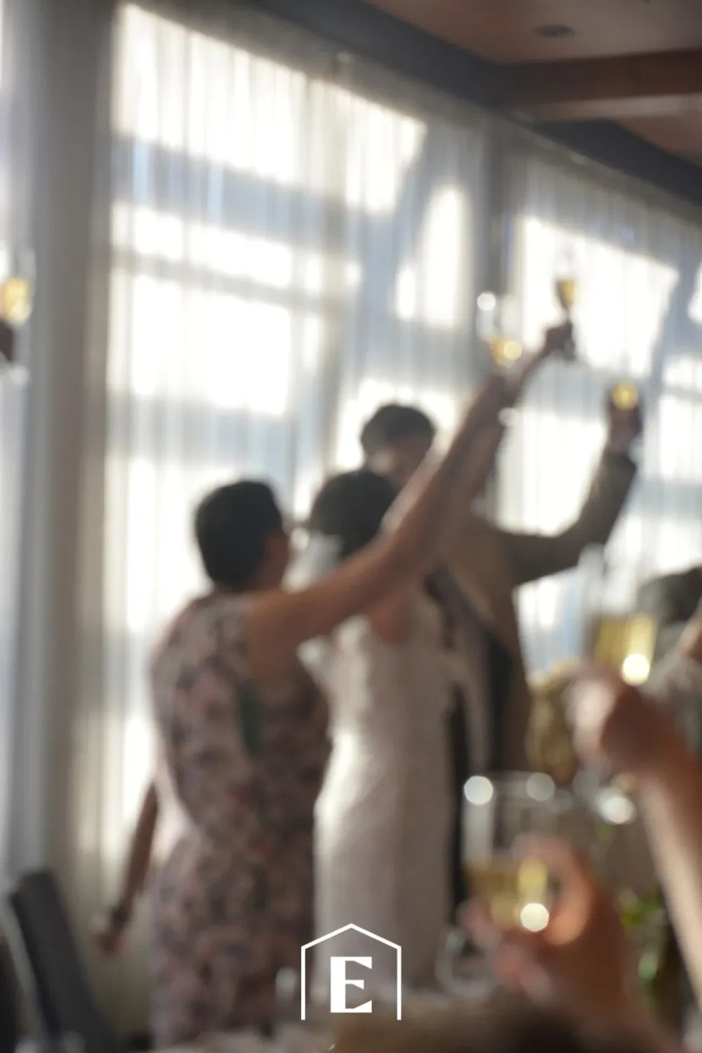 In this image of the Estrimont wedding venue, a couple is raising their champagne glasses in celebration, surrounded by a softly lit room. The bride, in a white gown, stands beside her partner, joined by another individual dressed in floral attire, all toasting to the occasion. Sunlight streams through large windows with sheer curtains, creating a warm and inviting atmosphere that enhances the elegance of the event. The out-of-focus foreground suggests a joyous wedding reception, capturing the essence of celebration and togetherness.