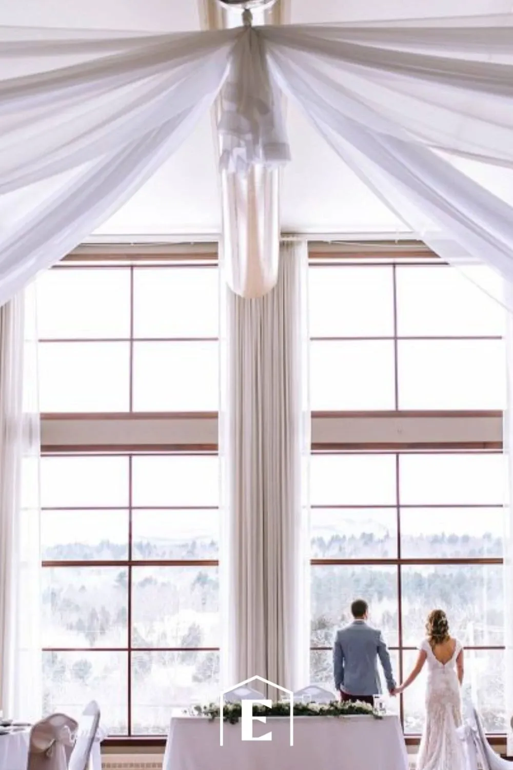 The image showcases a beautifully decorated wedding venue at Estrimont, under the category of Wedding Venues, part of the Mariage Elegant listing in Canada. The setting includes a spacious room with large windows that let in abundant natural light, offering a breathtaking view of the scenic landscape outside. Elegant white drapes hang from the ceiling, creating a sophisticated and romantic atmosphere. In the foreground, a couple stands together, gazing out the window, adding a personal and intimate touch to the venue's ambiance.