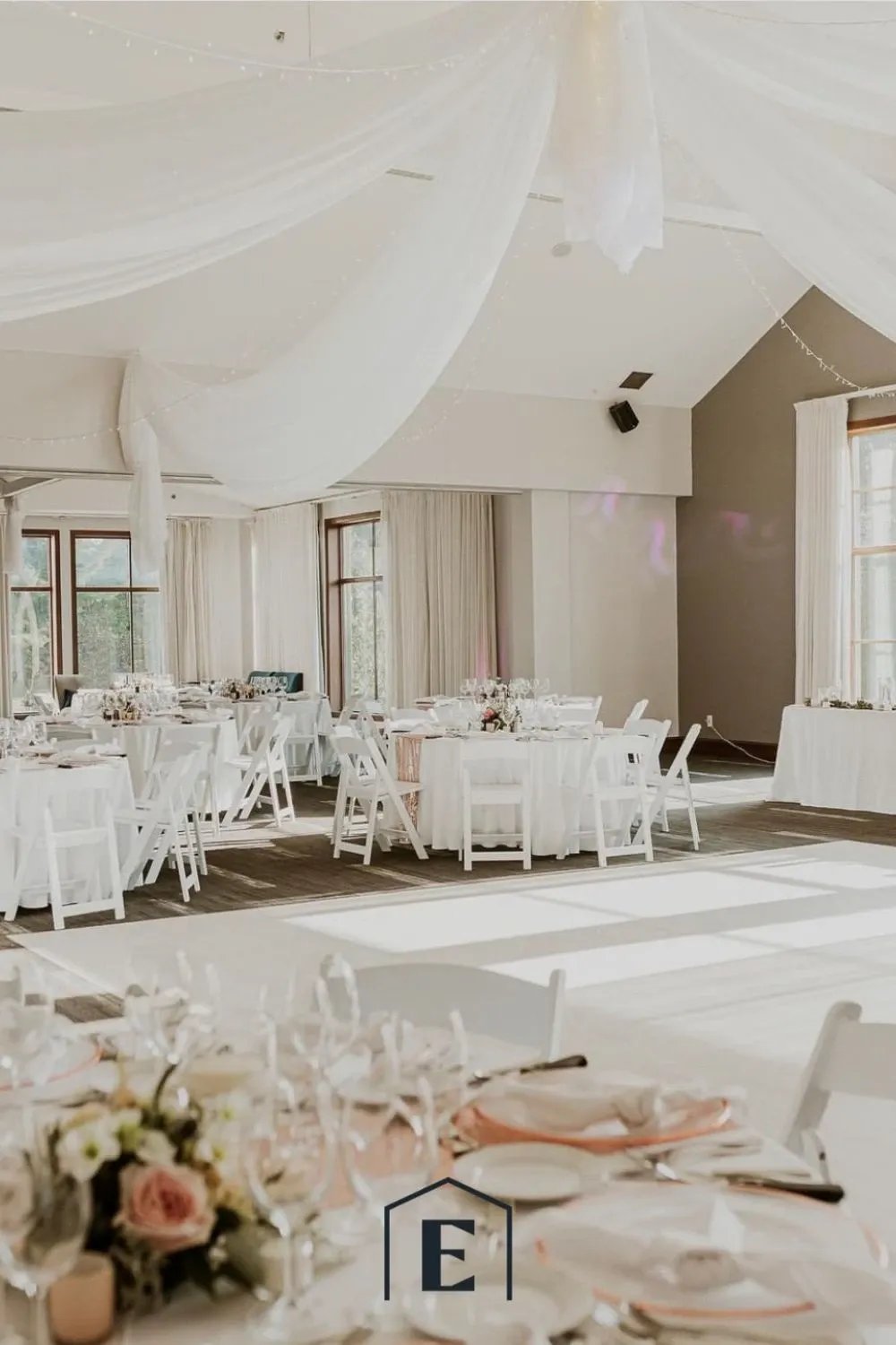 The image showcases an elegant wedding venue by Estrimont, categorized under Wedding Venues at Mariage Elegant, Canada. The spacious room features high ceilings with romantic, white draped fabric hanging gracefully. Round tables are elegantly set with white tablecloths, fine china, and blush pink accents, providing a sophisticated ambiance. Each table is adorned with delicate floral centerpieces. Large windows allow natural light to flood the space, enhancing the inviting atmosphere. This setting is perfect for a stylish wedding reception, offering both elegance and charm.