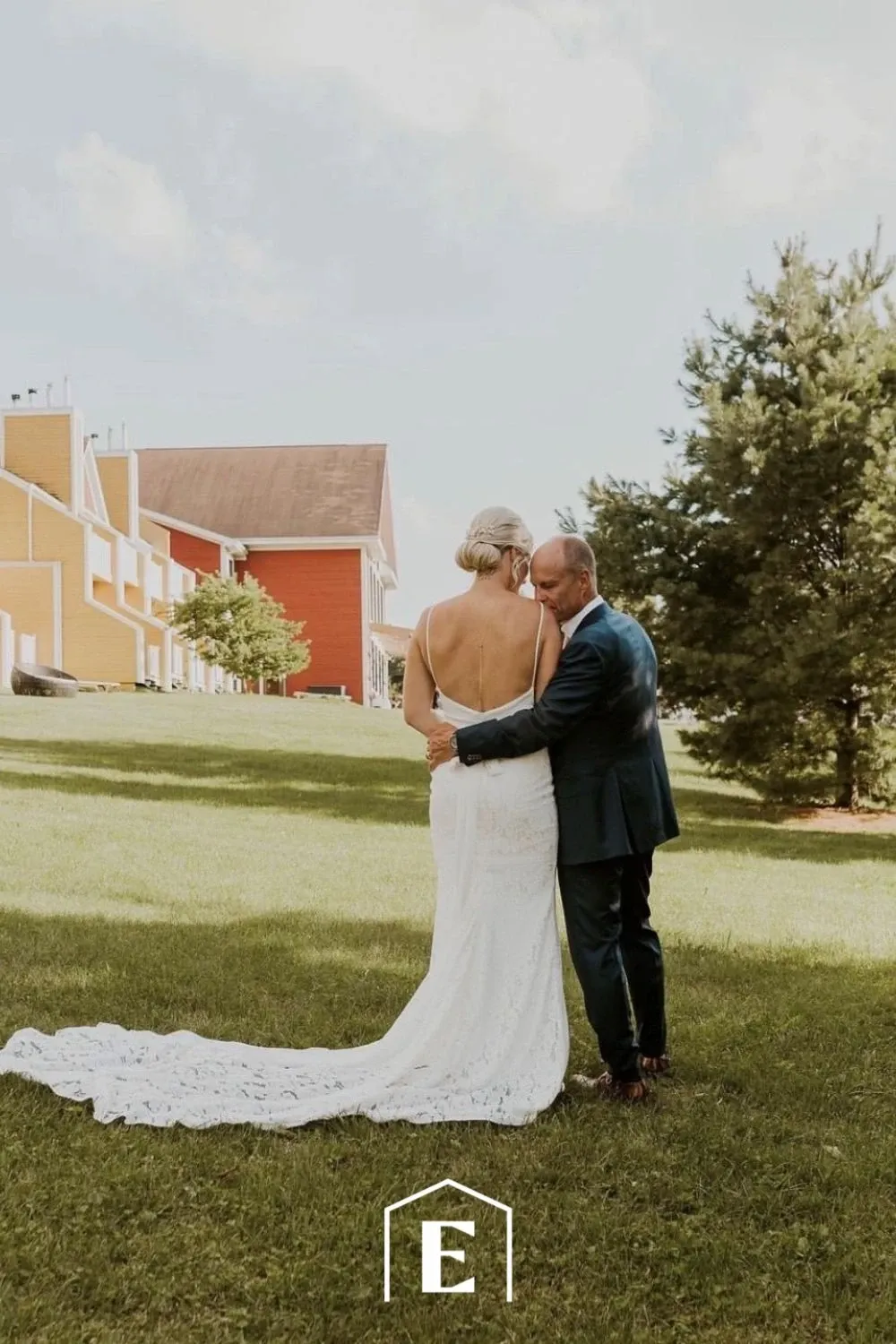 A newlywed couple is standing on a lush green lawn, embracing each other in front of Estrimont, a distinguished wedding venue in Canada. The backdrop showcases colorful buildings that complement the outdoor setting, with a mix of yellow and red structures providing a charming and inviting atmosphere. The bride is wearing an elegant white gown with intricate lace detailing and a long train, while the groom is dressed in a classic dark suit. The serene environment and thoughtful embrace highlight the romantic ambiance of this wedding venue. Estrimont offers an ideal setting for couples seeking a picturesque location for their special day.