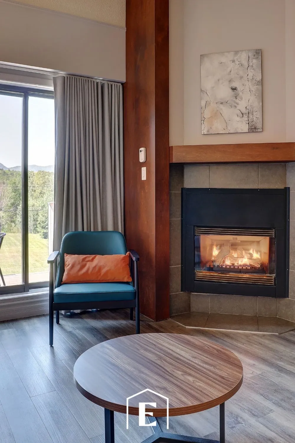The image showcases a cozy interior of Estrimont, a wedding venue listed under Mariage Elegant, Canada. It features a stylish lounge area with a plush armchair next to a glowing fireplace, creating a warm and inviting ambiance. A circular wooden coffee table is positioned in the foreground. Natural light streams through a large window adorned with soft gray curtains, offering a serene view of lush green trees outside. The setting exemplifies a tranquil and elegant atmosphere suitable for wedding events and gatherings.