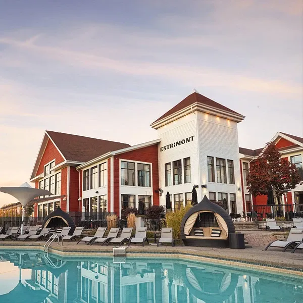 The image shows Estrimont, a wedding venue listed under Mariage Elegant in Canada. It features a charming, two-story building with a striking blend of red brick and white siding, highlighted by large windows. The venue is surrounded by a spacious outdoor area featuring a pristine swimming pool lined with lounge chairs and unique cabana structures, all set against a serene sky at dusk. This setting suggests a picturesque location ideal for hosting elegant wedding ceremonies and receptions.