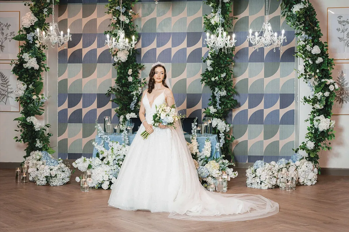 In this image, a bride dressed in a stunning white wedding gown holds a bouquet of white flowers. She stands in front of an elegant backdrop adorned with a geometric pattern, flanked by lush green foliage and blooming white flowers. Crystal chandeliers hang gracefully from above, casting a soft glow over the setting. The floor is covered in polished wood, adding a touch of warmth to the scene. This stylish setup, created by Di Event Sisters, highlights their expertise as top wedding planners in Canada for Mariage Elegant.