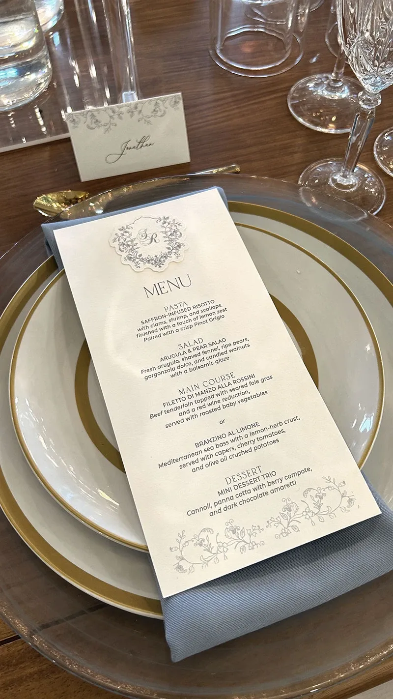 The image features an elegant wedding table setting arranged by Di Event Sisters, a vendor specializing in wedding planning under the category of Wedding Planners on the Mariage Elegant platform in Canada. A classic white and gold-edged dinner plate is placed on a glass charger, topped with a neatly folded gray-blue napkin. Centered on the napkin is a beautifully printed wedding menu with ornate designs at the top, listing gourmet food options. A delicate place card with the name 'Judith' and intricate floral patterns is positioned above the plate. Crystal-clear glassware and a polished wooden table further enhance the sophisticated and luxurious ambiance suitable for a wedding celebration.