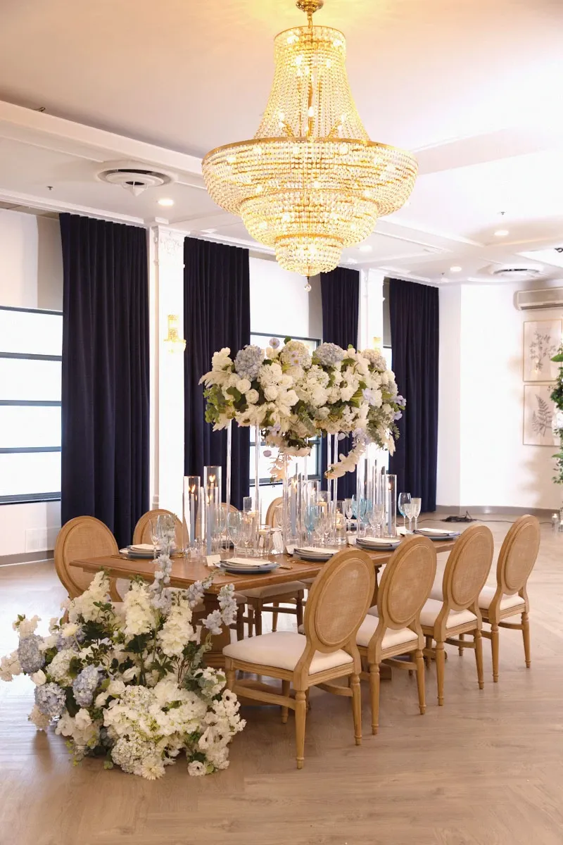 The image showcases an elegantly decorated wedding reception setup by Di Event Sisters in the Wedding Planners category. A long wooden table is beautifully arranged with a centerpiece featuring lush white and blue floral arrangements. The table is set with fine glassware and numerous tall, slender candles, contributing to a romantic ambiance. The chairs are upholstered in a soft beige fabric, complementing the overall sophisticated look. Above the table, a grand, sparkling chandelier casts a warm glow, enhancing the luxurious setting. The backdrop includes dark navy curtains, adding a touch of contrast to the bright decor. This luxurious setup reflects the meticulous planning capabilities of Di Event Sisters, perfect for a stylish and sophisticated wedding celebration.