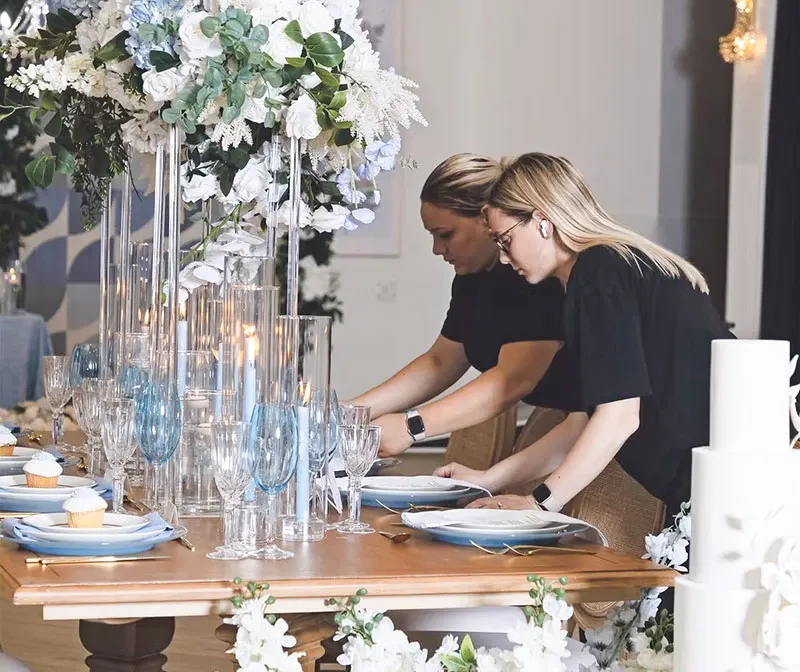 In this image, two women, likely from Di Event Sisters, are meticulously setting up a wedding table as part of their services in the wedding planners category. They are arranging elegant tableware, which includes blue-accented glassware and white plates, under a lavish floral centerpiece featuring white roses and lush greenery. The venue is adorned with sophisticated decor, including tall glass candle holders and delicately placed flowers, embodying a refined atmosphere perfect for an elegant wedding celebration.