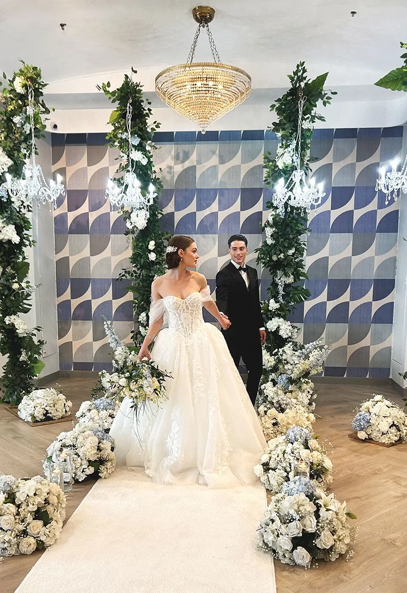 A bride and groom stand elegantly in a beautifully decorated wedding venue, highlighting the exceptional work of Di Event Sisters, a premier wedding planner in Canada. The bride wears a stunning, white, off-the-shoulder gown with intricate lace details, while the groom is dressed in a classic black tuxedo. They are surrounded by an opulent display of white and cream floral arrangements and lush greenery, which line the aisle and frame the couple. Overhead, crystal chandeliers and a unique geometric wall design add to the luxurious ambiance. This image exemplifies the sophisticated and meticulous wedding planning expertise offered by Di Event Sisters in the wedding planner category.