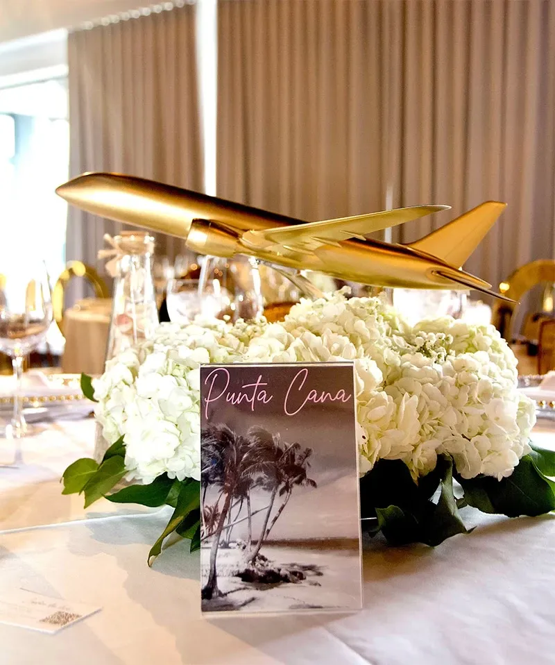 The image showcases a luxurious wedding table setup, curated by Di Event Sisters, a prominent wedding planner under the Mariage Elegant vendor listing in Canada. At the center, a golden airplane model gracefully rests atop a lush arrangement of white hydrangeas, symbolizing travel or a destination theme. In front of the floral display, a card inscribed with 'Punta Cana' and featuring a serene beach scene suggests a tropical destination or theme for the event. The background features neatly arranged dining tables with wine glasses, exuding a sophisticated and elegant ambiance, ideal for weddings.