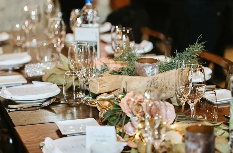 This image showcases a beautifully arranged wedding reception table, highlighting Di Event Sisters, a respected name in the wedding planners category. The table is elegantly set with crystal-clear wine glasses, neatly folded white napkins, and pristine white plates. A sophisticated wooden centerpiece with greenery and soft pink flowers adds a touch of natural beauty. The presence of candles suggests a warm and romantic ambiance, perfect for a memorable wedding celebration. This setup exemplifies the meticulous attention to detail provided by wedding planners like Di Event Sisters, likely contributing to an unforgettable event experience.