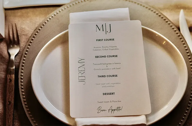 The image features an elegant wedding table setting with a neatly arranged dinner menu placed on a white plate over a soft brown woven charger plate. The menu lists courses including risotto, bolognese, and desserts, suggesting a wedding reception planned by Di Event Sisters, categorized under Wedding Planners at Mariage Elegant, Canada. The setup exudes sophistication with its neutral tones and fine dining arrangement, reflecting meticulous attention to detail typical of professional wedding planning.