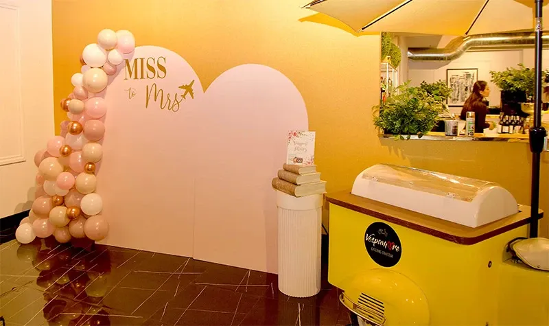 This image captures a whimsical setup by Di Event Sisters, a vendor in the Wedding Planners category at Mariage Elegant, Canada. The scene features a pastel pink heart-shaped backdrop with 'Miss to Mrs' elegantly written on it, accompanied by a cluster of pink and white balloons. To the right, a charming yellow cart with a decorative umbrella adds a playful touch, while a staff member interacts behind a counter adorned with green foliage. The setup exudes a joyful, celebratory ambiance, perfect for a bridal shower or pre-wedding event.