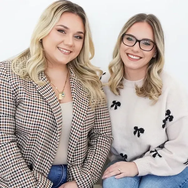 The image features two smiling women who are part of the Di Event Sisters, a wedding planning vendor in the Wedding Planners category for Mariage Elegant, Canada. The woman on the left is wearing a plaid blazer and a beige top, accessorized with a layered necklace. The woman on the right is wearing glasses, a light-colored sweater with black bow designs, and a ring on her finger. They both have long, blonde hair and are seated against a plain white background, exuding a professional and approachable demeanor, suitable for wedding planning services.