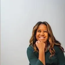 The image shows a woman with long, wavy hair smiling confidently while seated, wearing a dark green blouse. She has a thoughtful pose with her chin resting on her hand against a neutral, light background. This image represents Conception Ezzahra, a vendor specializing in wedding planning services, featured on the Mariage Elegant website in Canada. The professional demeanor of the individual suggests expertise in coordinating elegant wedding events, making her a valuable asset to couples seeking assistance for their special day.