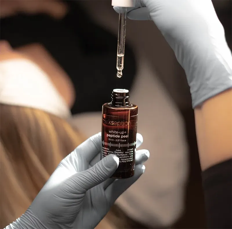In this image related to wedding beauty by Clinique Âme, a person wearing blue gloves is holding a small brown bottle labeled 'White-Up Peptide Peel.' The bottle is being used with a dropper, dispensing a liquid, suggesting a skincare or beauty treatment suitable for brides. In the background, there is a blurry figure lying down, typical of a spa or beauty clinic setting, indicating a focus on personal care and wedding preparation services offered by the vendor.