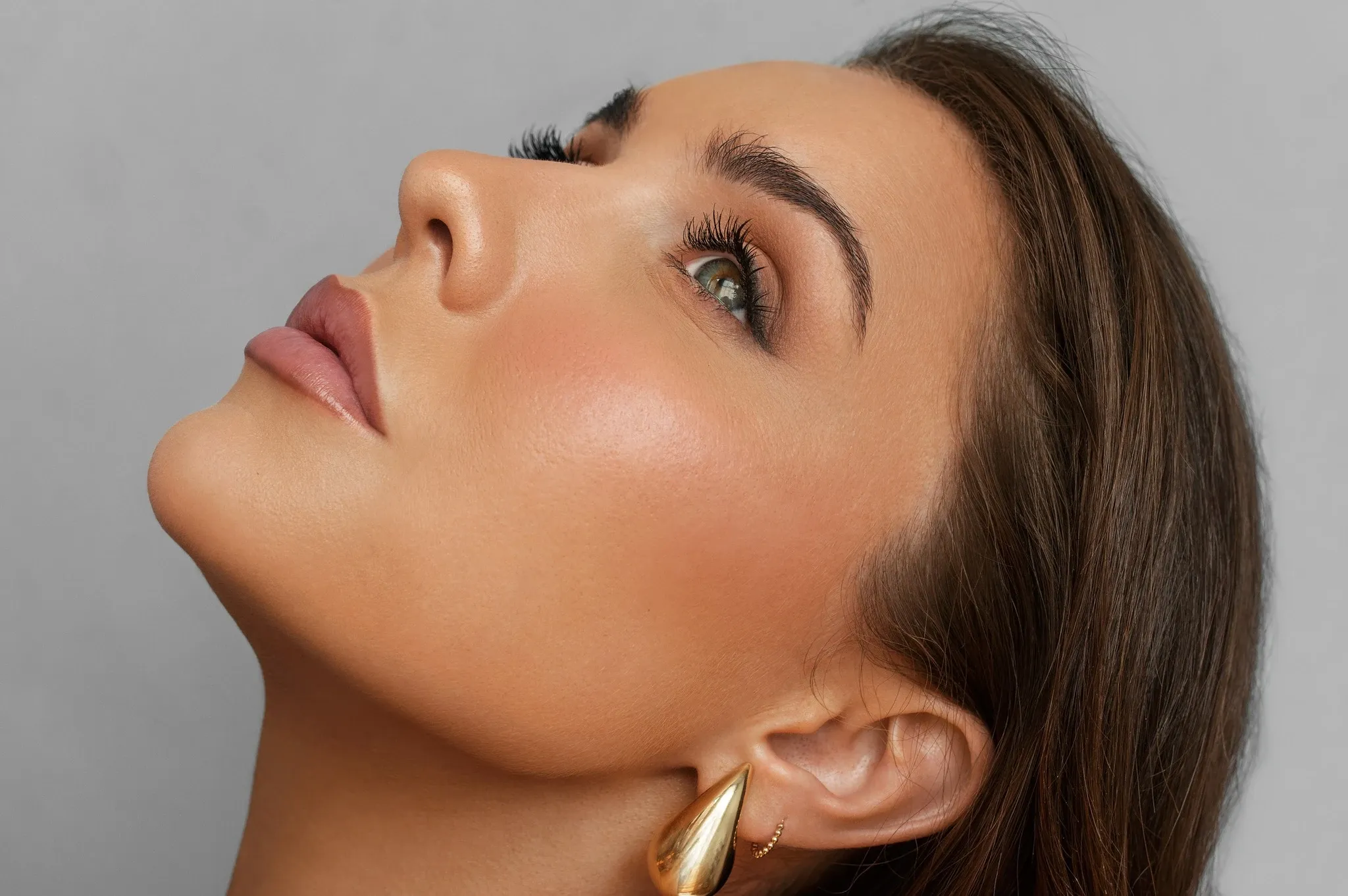 In this close-up image, a woman gazes upwards, showcasing a flawless bridal makeup look. Her skin is smooth and radiant, with expertly applied foundation, soft blush, and perfectly contoured features. Her eye makeup features subtle brown and gold tones, emphasizing her natural beauty. She wears elegant, gold teardrop earrings that add a touch of sophistication. This image is associated with Clinique Âme, a vendor in the Wedding Beauty category, highlighting their expertise in creating stunning bridal looks for wedding events.