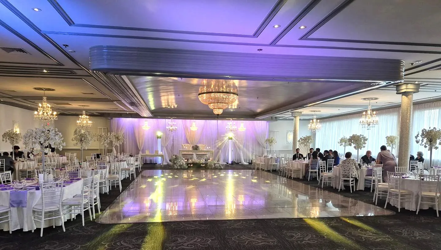 The image showcases an elegant wedding venue at Buffet Marina, a featured vendor in the Wedding Venues category on Mariage Elegant, Canada. The spacious room is beautifully decorated with white and purple floral arrangements on tables set with crisp white linens. A grand chandelier hangs from the ceiling, casting a warm glow over the polished dance floor. The venue is filled with guests seated on white chairs, enjoying the sophisticated atmosphere. The lighting design includes purple uplighting that enhances the ambiance, highlighting the draped white backdrop behind the head table. This sophisticated setting reflects the high standards of Buffet Marina as a premier wedding venue choice.