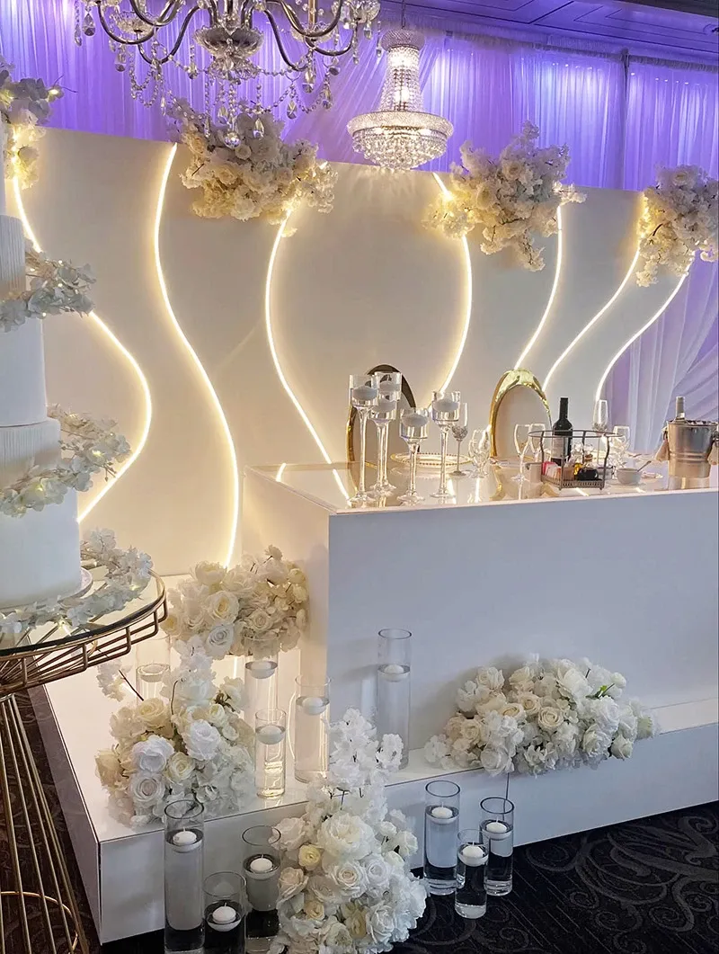 The image showcases a beautifully decorated wedding venue by Buffet Marina, highlighting the sophistication associated with top-tier wedding venues in Canada. The setting features an elegant white bar adorned with cascading white floral arrangements and sleek glass candle holders lined with candles. A backdrop with a modern, wavy design and subtle lighting adds a luxurious ambiance. Overhead, a stunning chandelier enhances the sophisticated atmosphere, setting the scene for a memorable wedding reception. This setup exemplifies the professional and elegant style expected from wedding venues partnered with Mariage Elegant.