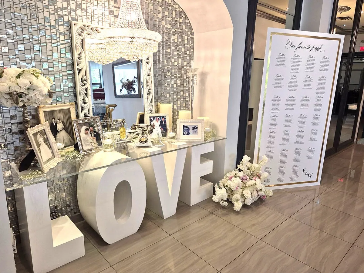 This image showcases an elegant decor setup at Buffet Marina, a featured wedding venue in the Mariage Elegant vendor listing. The scene includes a reflective mosaic-tiled wall adorned with a stunning, ornate chandelier overhead. In front of the wall, a glass-topped table is stylishly decorated with framed photographs, white candles, and floral arrangements. The large white block letters spelling 'LOVE' add a romantic touch to the display. To the right, a seating chart on an easel lists guests' names, indicating meticulous wedding planning and reception organization. The overall ambiance is sophisticated and inviting, perfect for hosting a memorable wedding celebration.