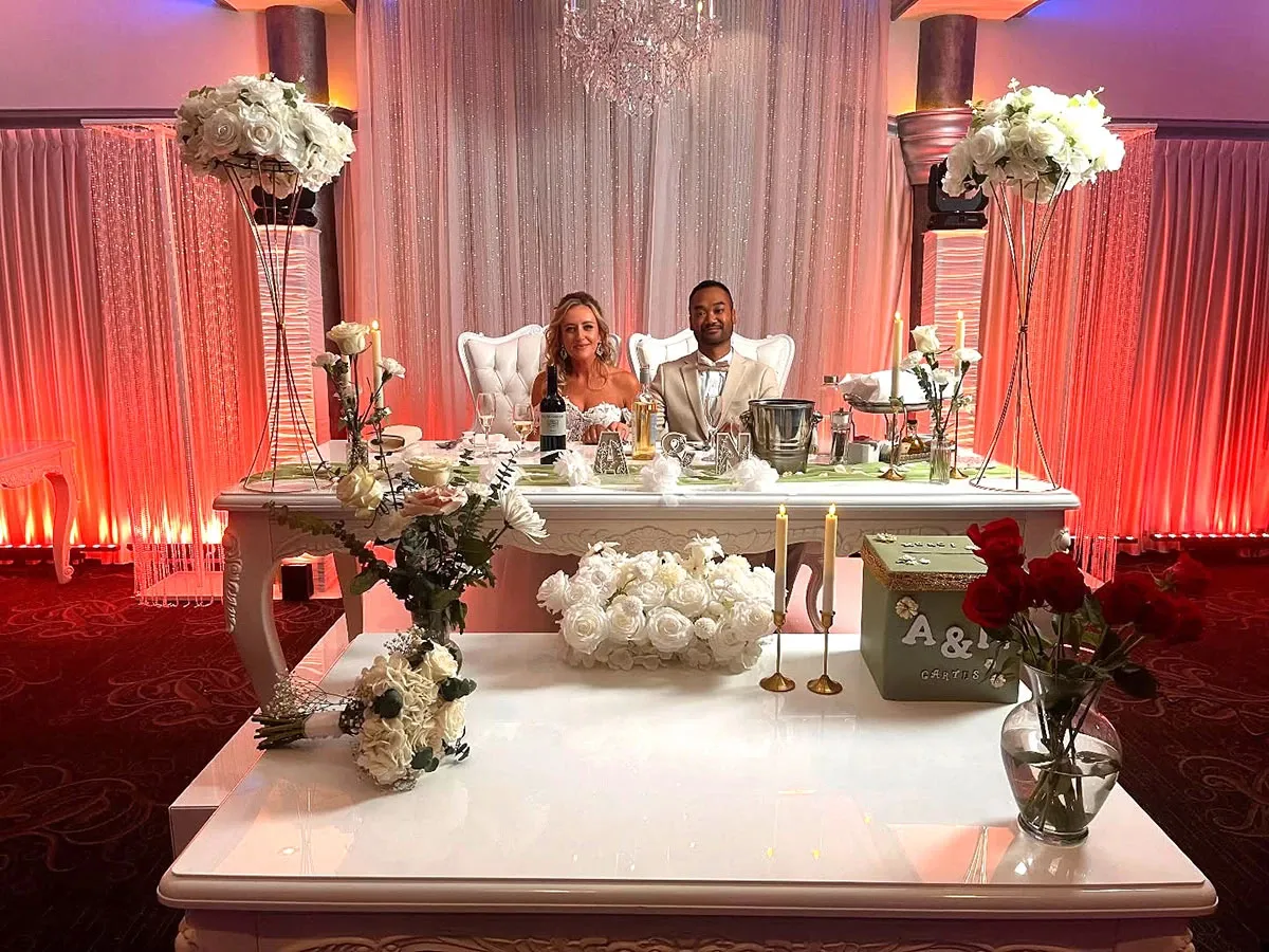 The image features a beautifully decorated wedding venue by Buffet Marina, showcased within the wedding venues category on Mariage Elegant, Canada. At the center is a long, elegant table adorned with white flowers, candles, and sparkling glassware. A couple sits behind the table, smiling, in a setting illuminated by warm, ambient red and gold lighting that enhances the romantic atmosphere. Tall floral arrangements on either side add to the sophisticated decor. This exquisite setup reflects Buffet Marina's commitment to creating stunning and memorable wedding experiences.