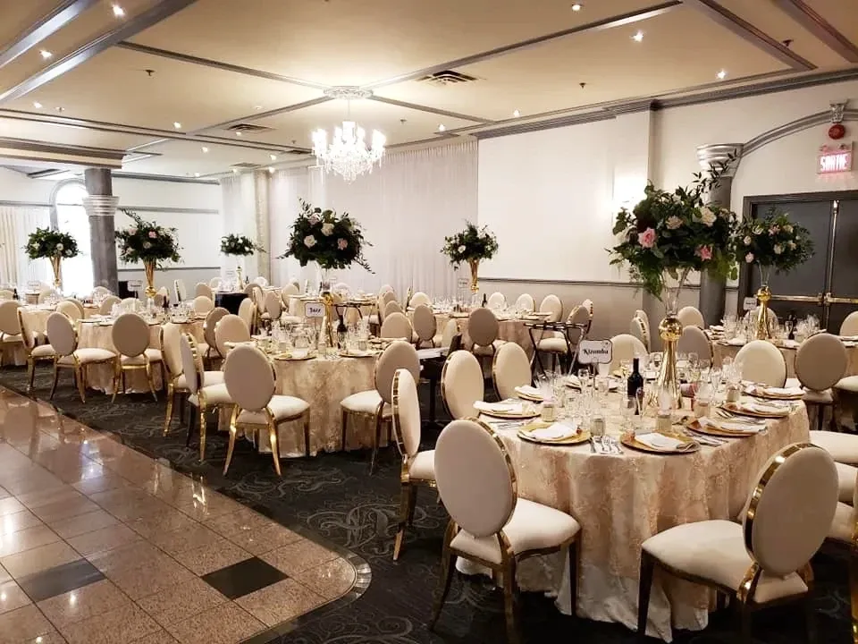 The image shows an elegant wedding venue provided by Buffet Marina, categorized under Wedding Venues on Mariage Elegant, Canada. The spacious room is beautifully decorated with round tables covered in luxurious beige tablecloths, each adorned with stunning floral centerpieces consisting of white and pink roses, and lush greenery. The room features sophisticated cream-colored chairs with gold trim, complementing the ornate setting. A sparkling chandelier hangs from the ceiling, adding a touch of glamour and elegance to the venue. The soft ambient lighting and the tastefully neutral decor create a refined and inviting atmosphere, ideal for a memorable wedding celebration.