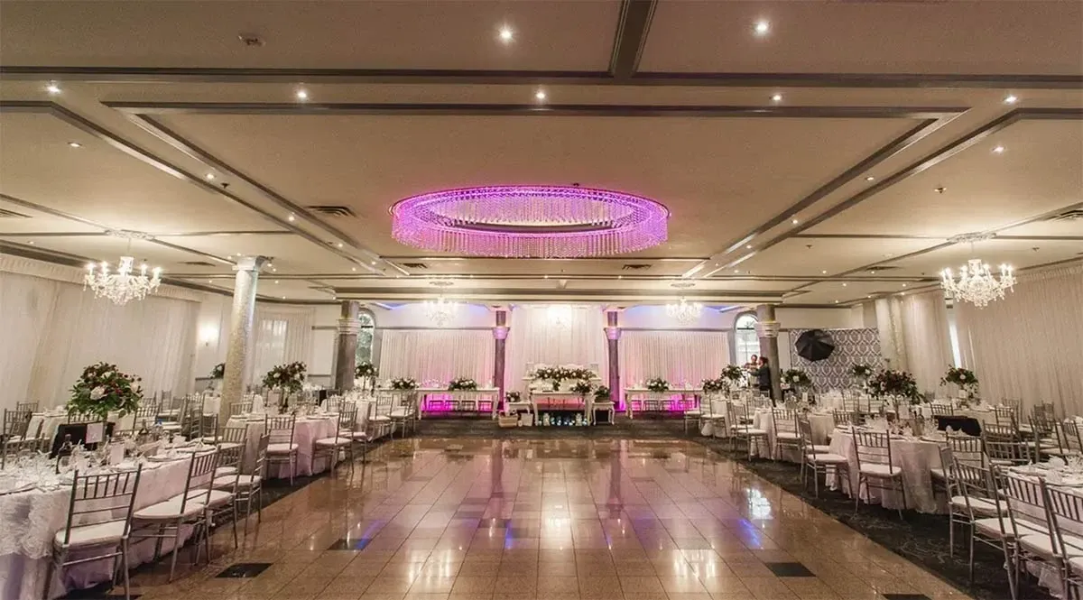 The image shows an elegant wedding reception setup at Buffet Marina, a wedding venue featured on Mariage Elegant, Canada. The spacious hall is adorned with round tables covered in white tablecloths, each surrounded by silver Chiavari chairs. The tables are elegantly set with fine silverware and feature sophisticated floral centerpieces. A large, modern chandelier with pink lighting hangs from the ceiling, adding a touch of glamour to the ambiance. The room is softly lit with additional chandeliers and recessed lighting, creating a warm and inviting atmosphere. In the background, a beautifully decorated head table is positioned against a tastefully draped backdrop, enhancing the venue's luxurious appeal.