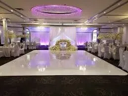 The image shows an elegant wedding venue provided by Buffet Marina, categorized under Wedding Venues, on the Mariage Elegant website in Canada. The spacious interior features a glossy white dance floor surrounded by round tables draped in white tablecloths and adorned with floral centerpieces. Soft purple lighting adds a romantic ambiance, while a large, illuminated chandelier hangs from the ceiling, creating a focal point above the dance floor. In the background, a beautifully decorated arch and floral arrangement highlight the space for the ceremony or speeches, offering a sophisticated setting for a memorable wedding celebration.