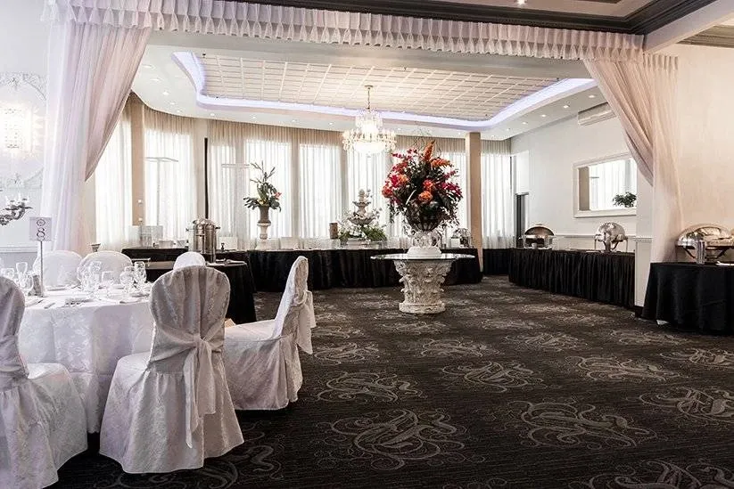 The image features an elegantly decorated wedding venue by Buffet Marina, a vendor listed under the Wedding Venues category on Mariage Elegant, Canada. The spacious room is adorned with lavish white drapes and an exquisite chandelier hanging from the ceiling, casting a warm glow over the setting. Round tables are neatly arranged, showcasing pristine white tablecloths and covered chairs, creating a sophisticated atmosphere. In the corner, a grand floral arrangement with vibrant red and pink flowers stands atop a pedestal, adding a touch of color and elegance. The venue exudes a sense of luxury, ideal for an elegant wedding celebration.