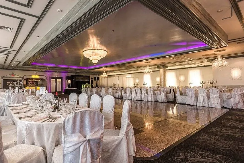 The image showcases an elegantly set up wedding venue offered by Buffet Marina, a premier vendor listed under wedding venues on Mariage Elegant, Canada. The spacious hall features a gleaming dance floor at the center, surrounded by round tables adorned with white tablecloths and neatly arranged chairs. Each table is decorated with sparkling glassware and neatly folded napkins, creating a sophisticated ambiance. A stunning chandelier hangs from the ceiling, adding a touch of glamour, while purple accent lighting enhances the romantic atmosphere. The venue is perfect for hosting large wedding receptions, combining elegance with functionality.