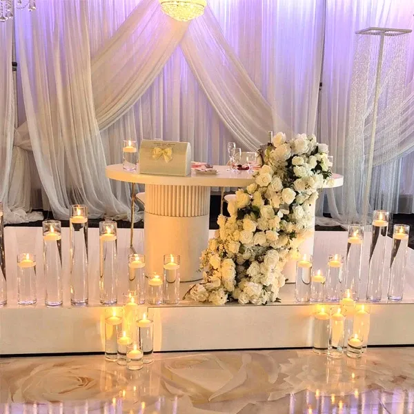 The image showcases a beautifully decorated wedding venue at Buffet Marina, a vendor listed under Wedding Venues on Mariage Elegant, Canada. The setting features a round table adorned with an elegant floral arrangement of white roses cascading gracefully. Numerous glass candles are artfully arranged around the table, casting a warm and inviting glow. The backdrop consists of sheer, flowing white drapes that contribute to the venue's sophisticated ambiance, perfect for a romantic wedding celebration. The setting captures the essence of elegance and style, making it an ideal choice for couples seeking a luxurious wedding venue.